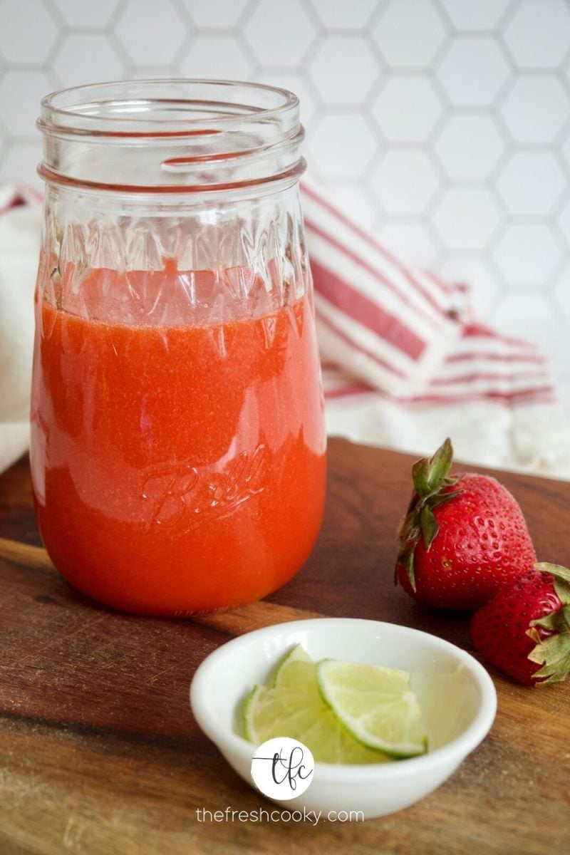 A mason jar of fresh strawberry syrup on a cutting board with strawberries and lime by the syrup.