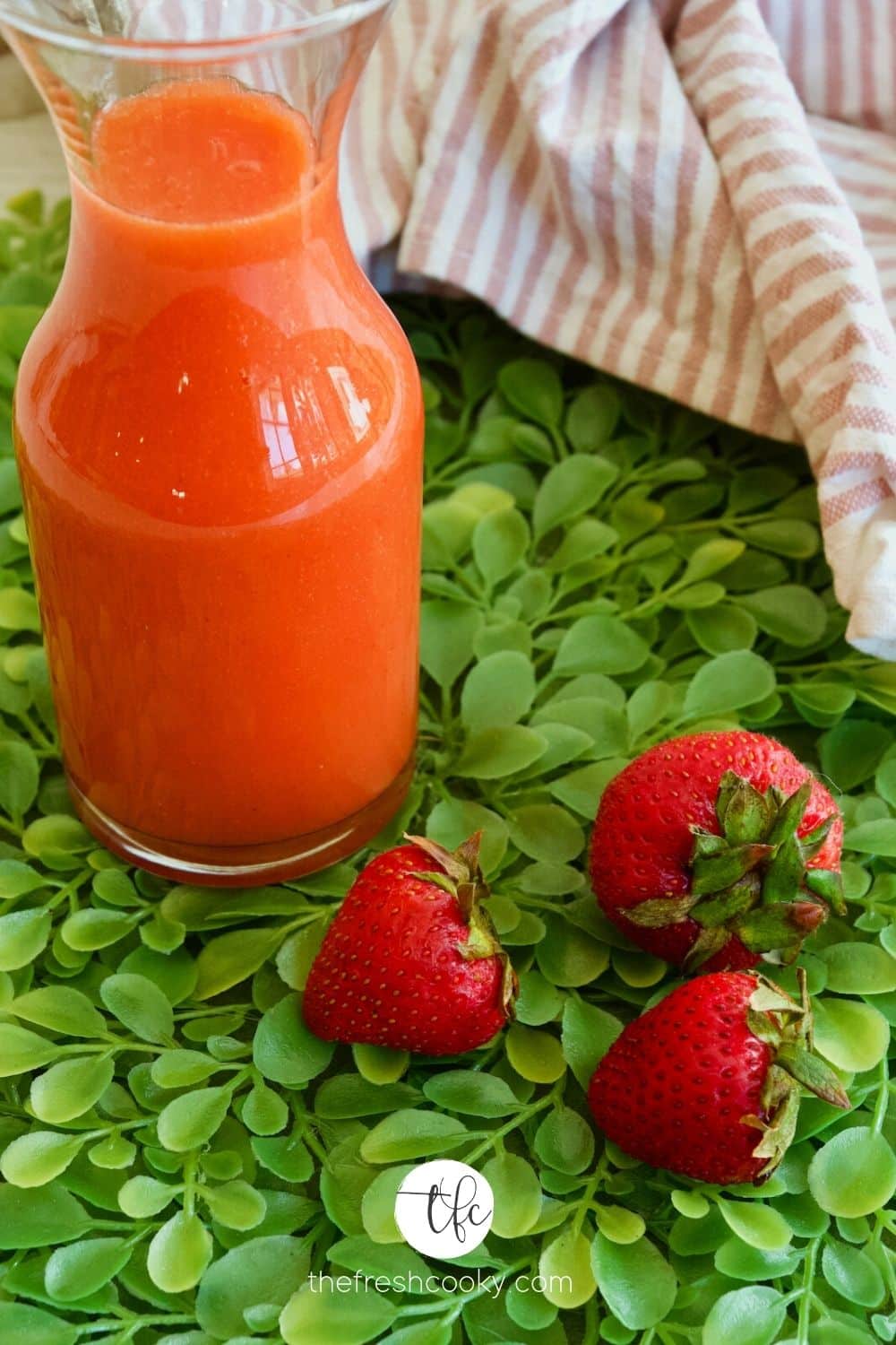 Carafe filled with bright red strawberry syrup on a bright green plant placemat with three fresh strawberries.