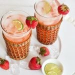 Two tall glasses with wicker jackets, filled with pink strawberry gin and tonic cocktail, garnished with limes and a strawberry.