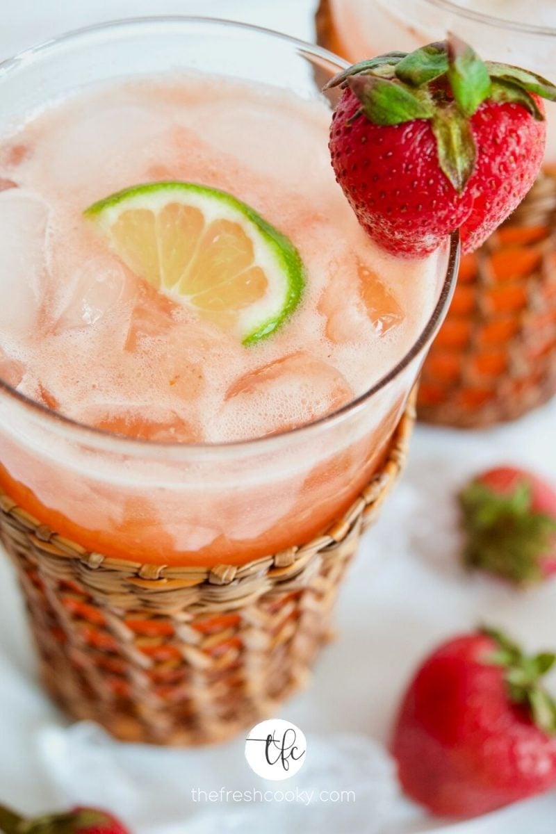 CLose up image of strawberry gin cocktail with wedge of lime and strawberry in a tall wicker jacketed glass.