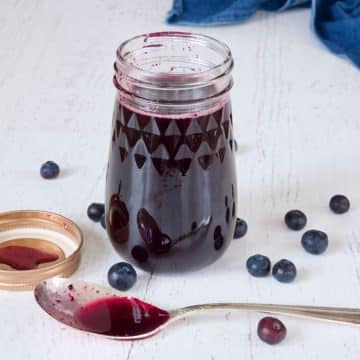 Image of jar of blueberry simple syrup with spoon in front and fresh blueberries laying about.