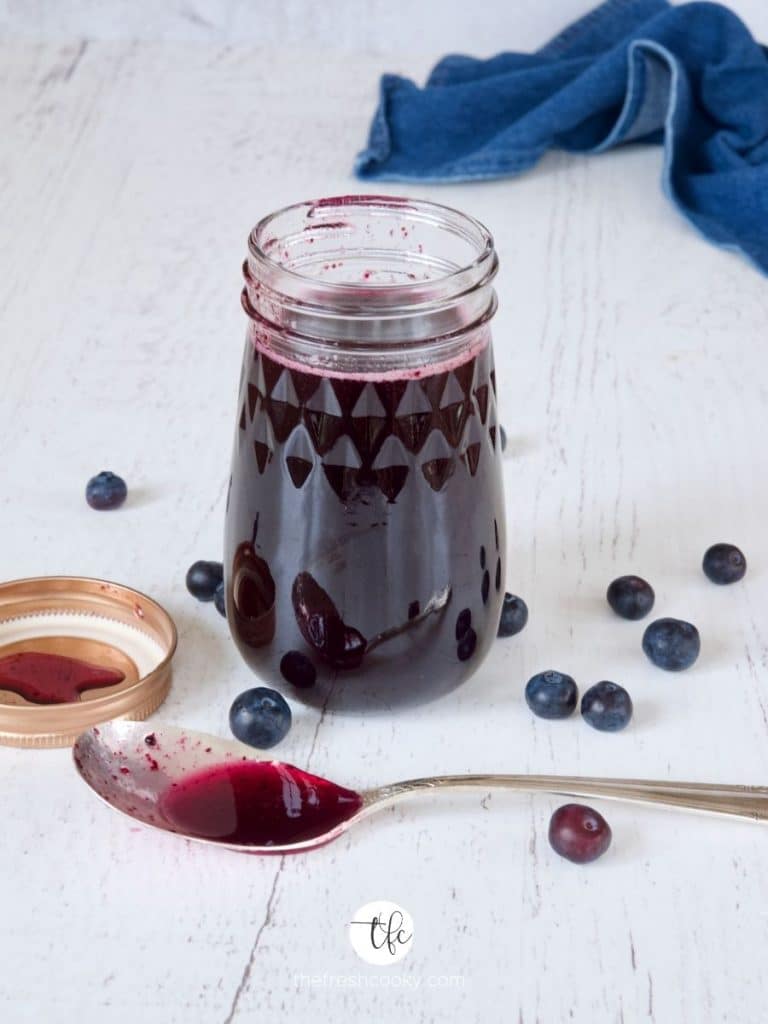 Image of jar of blueberry simple syrup with spoon in front and fresh blueberries laying about.