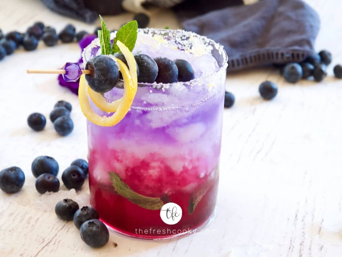 Blueberry Gin Cocktail with skeweered blueberries, lemon twist and blueberries laying around.
