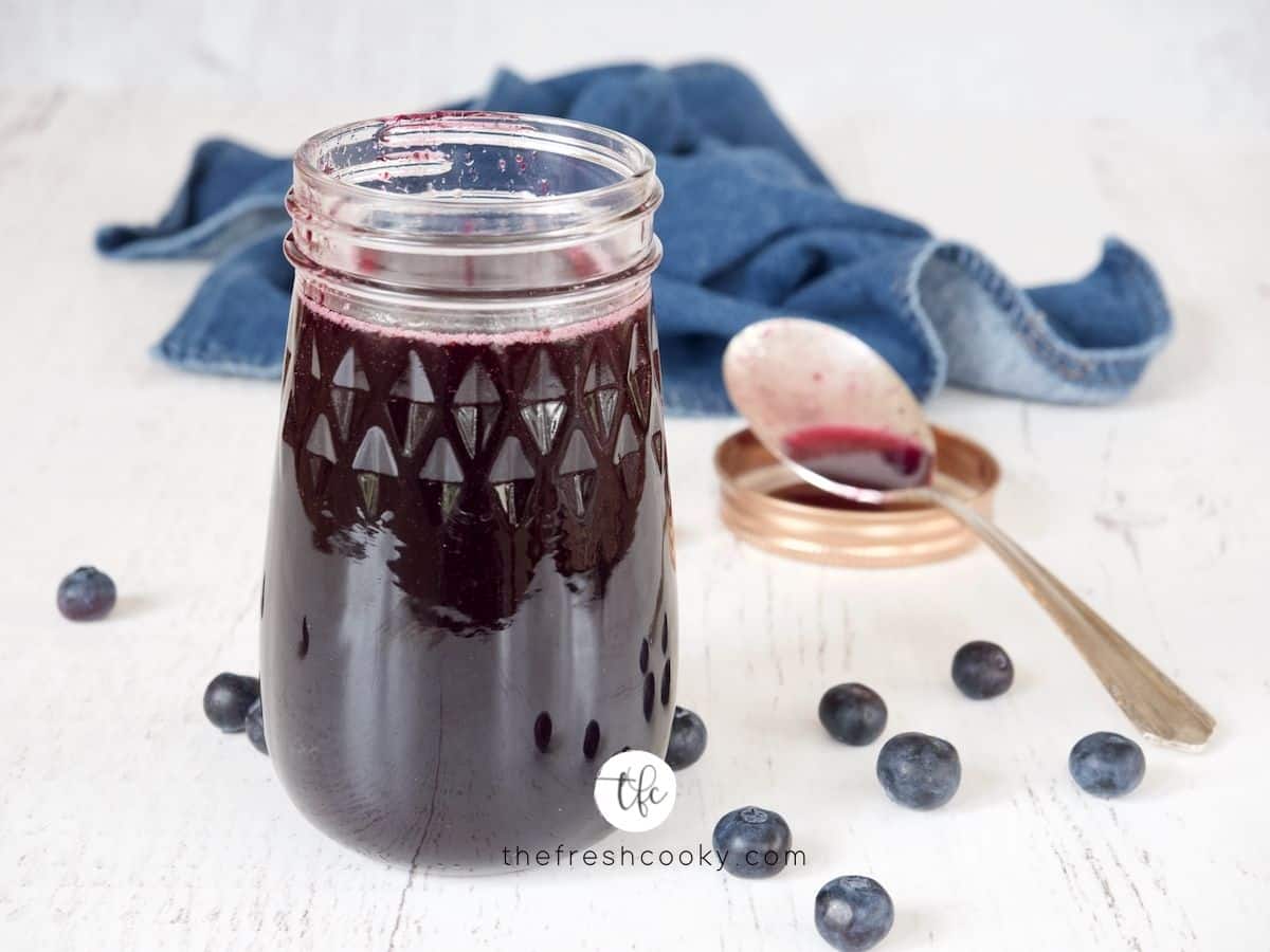 FB Image of Blueberry Simple Syrup on white backgroudn with fresh blueberries strewn about and a spoon with some syrup in it in the background.