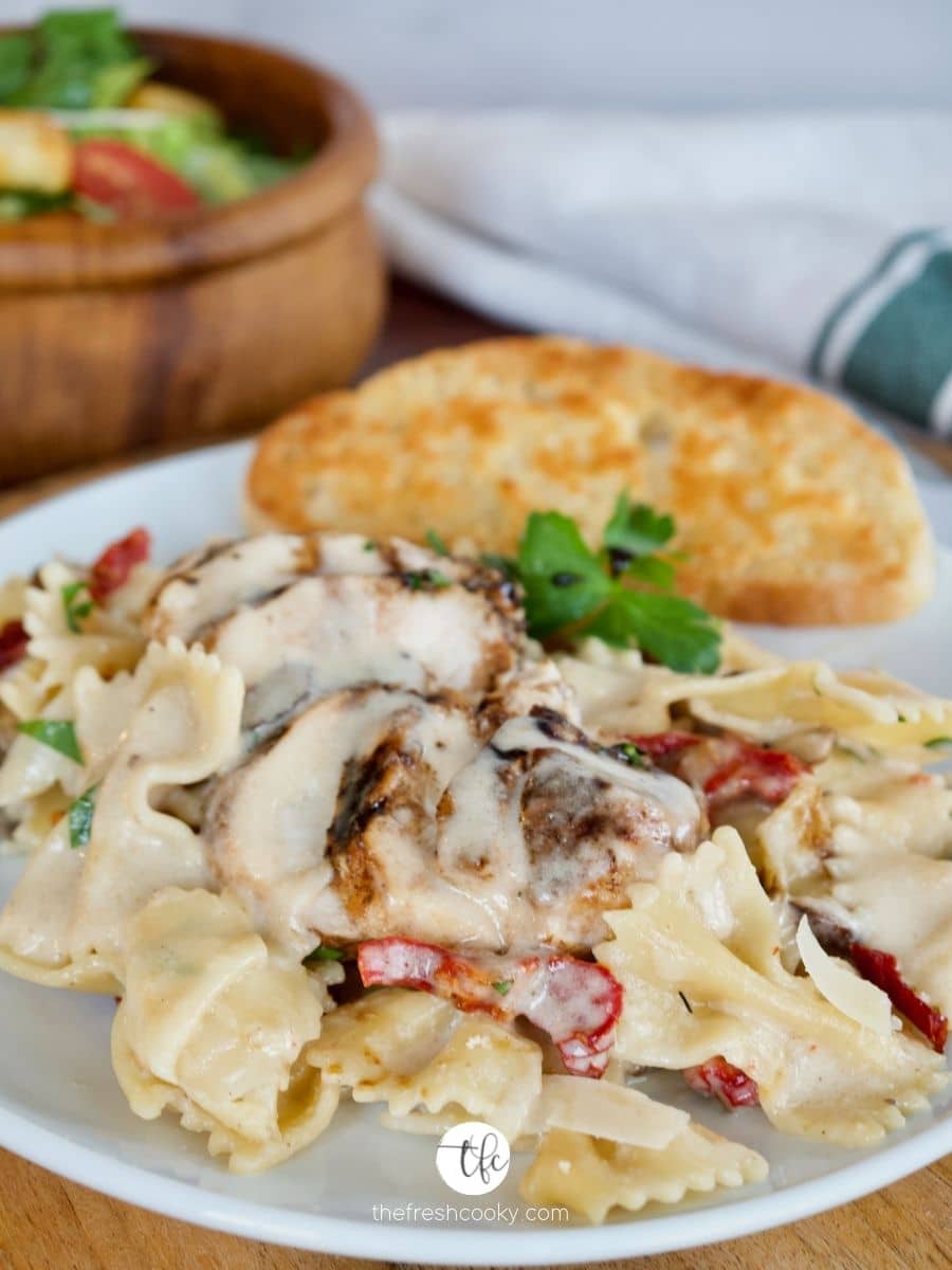 Creamy Bow Tie Pasta on white plate with garlic toast and a salad in background.