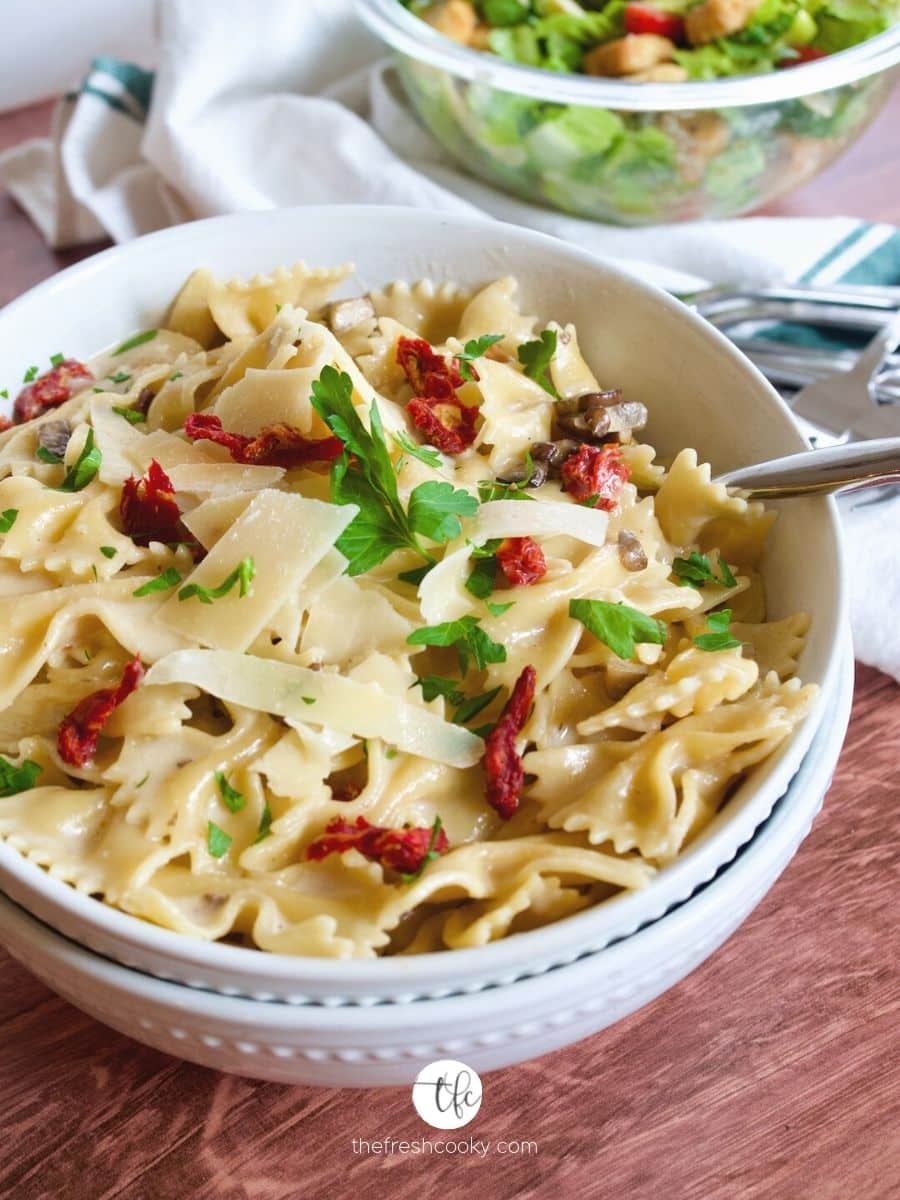 creamy bow tie pasta recipe with sun-dried tomatoes and mushrooms in large bowls with spoon.