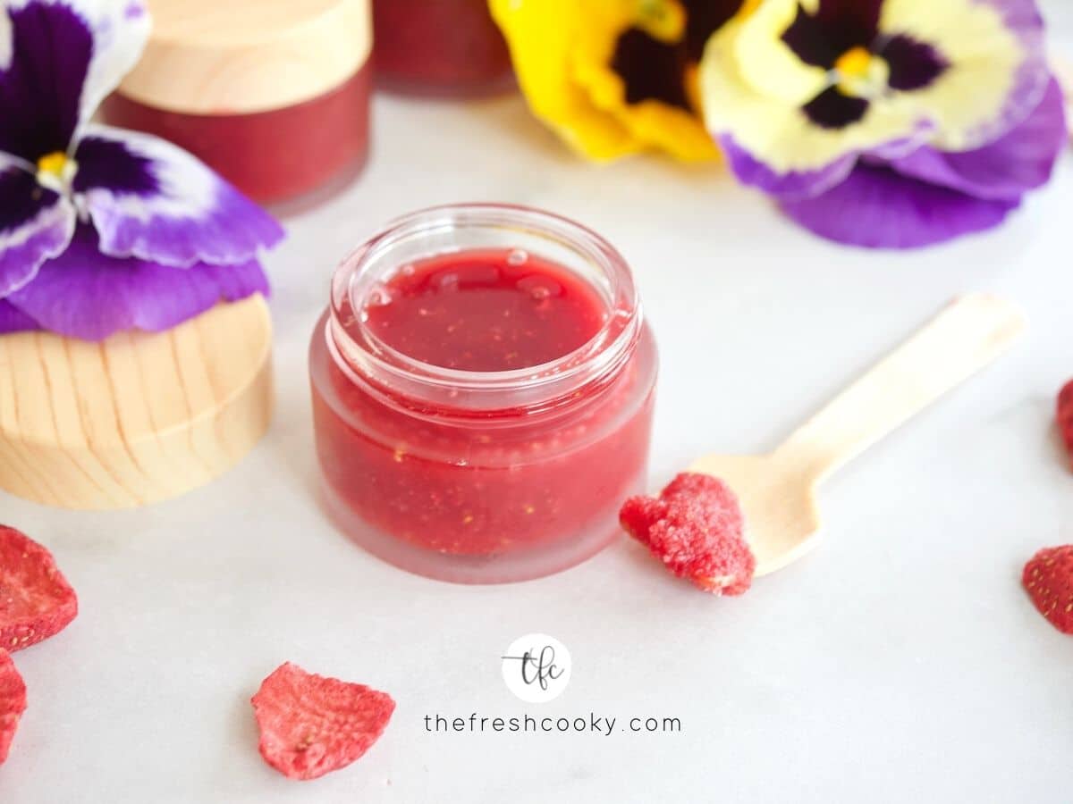 FB image for DIY Lip Scrub with strawberries, small glass jar filled with bright pink strawberry lip scrub with small spatula with a little on it.