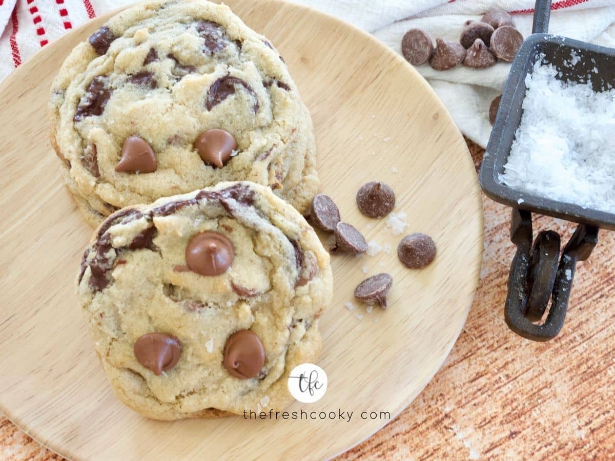 Best chewy gf chocolate chip cookies on wood grain plate with a small bowl of flaky sea salt nearby.