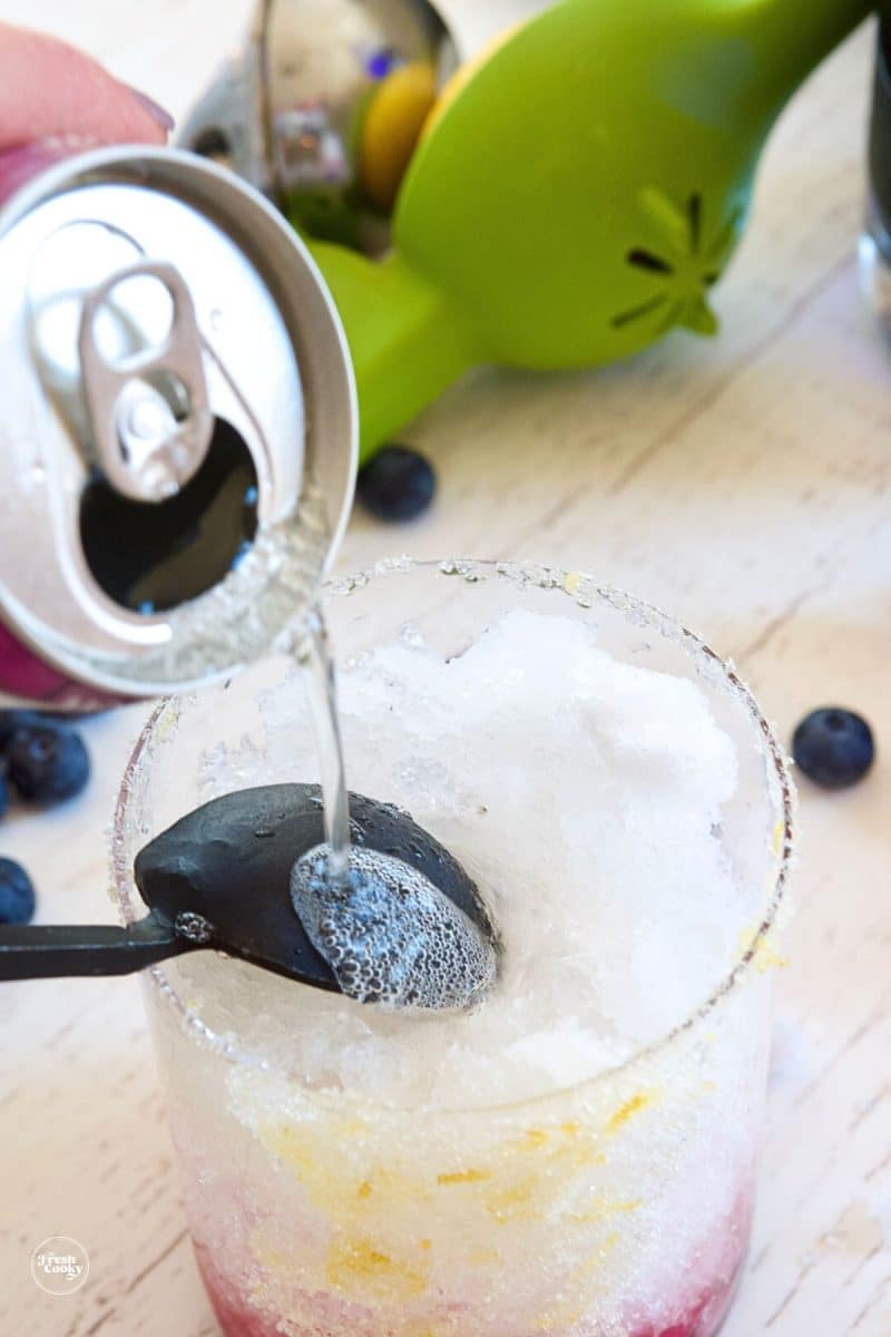 Pouring ginger beer over the back of a spoon to float on the blueberry syrup layer. 