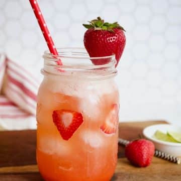 Gorgeous shot of strawberry refresher drink on wooden cutting board with pink straw and a strawberry garnish with limes and strawberries in the background.