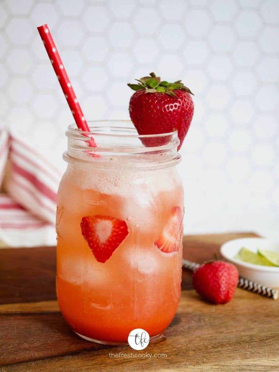 Gorgeous shot of strawberry refresher drink on wooden cutting board with pink straw and a strawberry garnish with limes and strawberries in the background.
