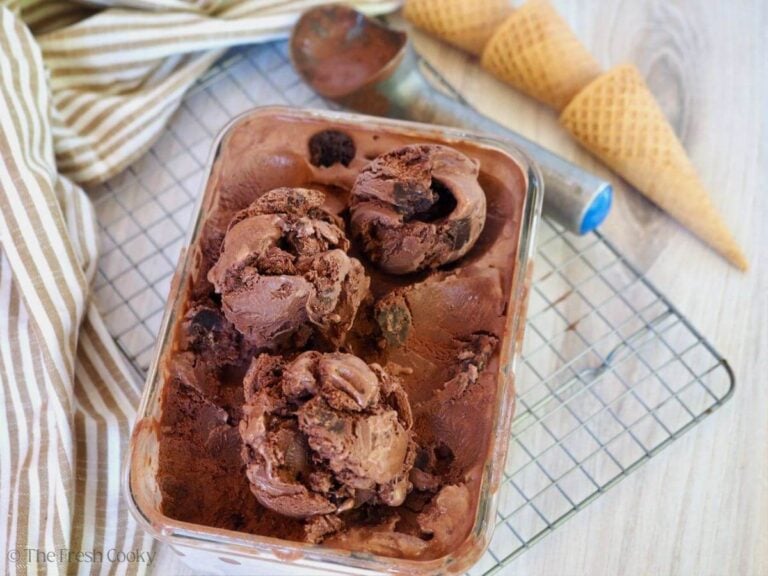 Three scoops of chocolate brownie ice cream in a container with sugar cones and a scoop nearby.