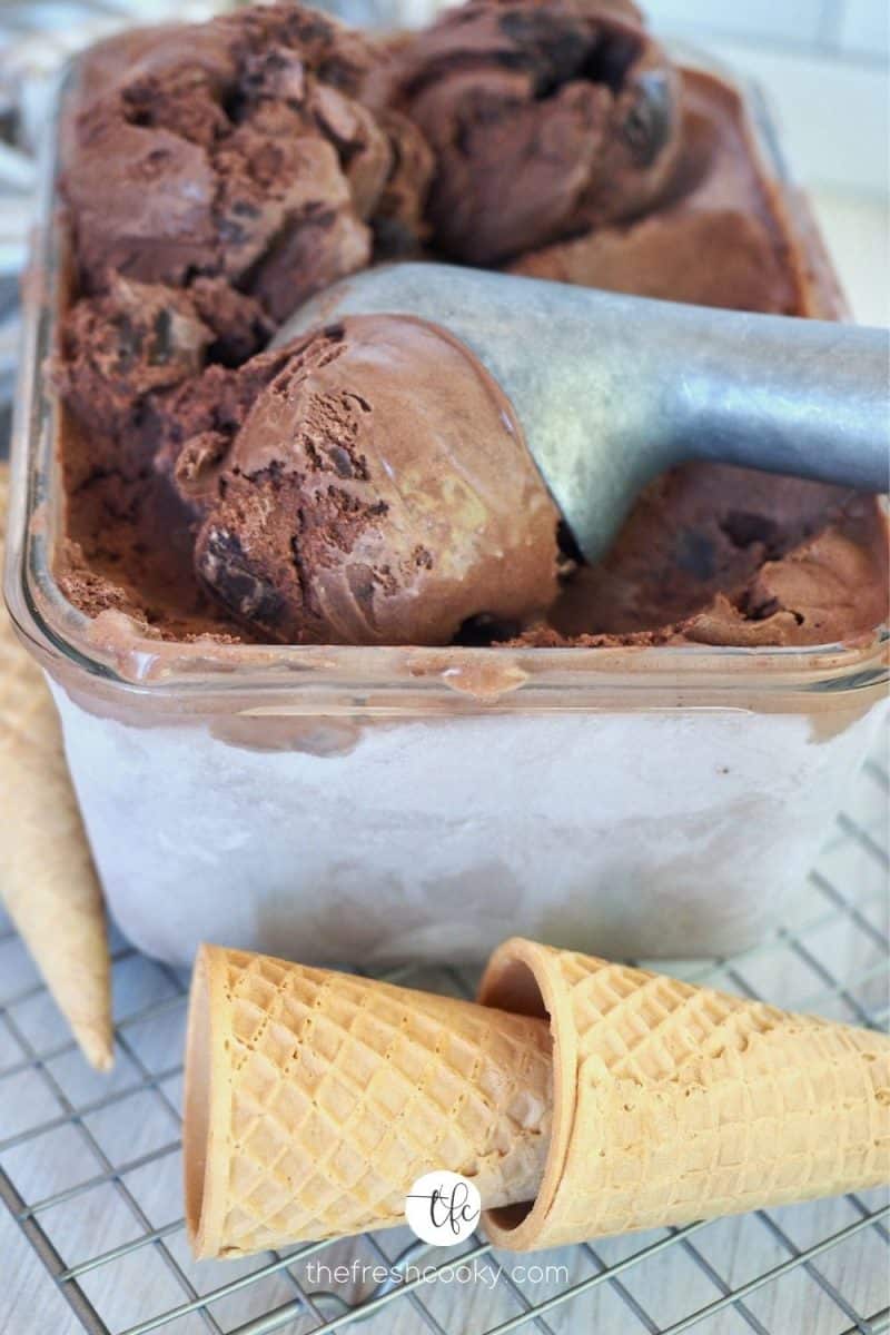 Frozen rectangler container with ice cream scoop scooping large creamy scoop of Chocolate Brownie Fudge Ice Cream with sugar cones in front.