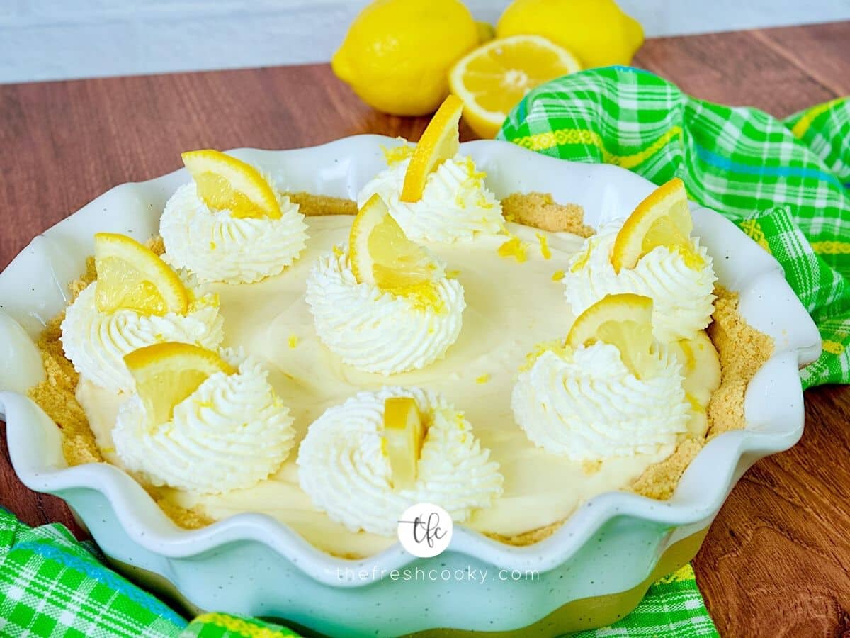 Lemon cream pie FB photo, with whipped cream dollups, decorated with a wedge of lemon with lemons in the background.