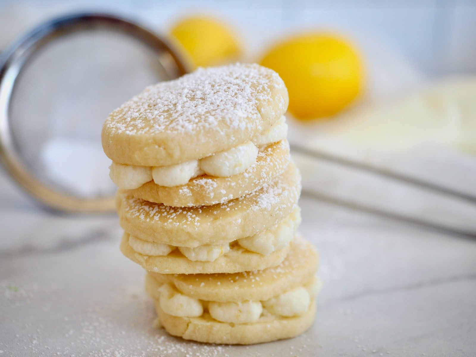 Lemon sandwich cookies three stacked on top of the other, with fresh lemons in background and seive with powdered sugar.