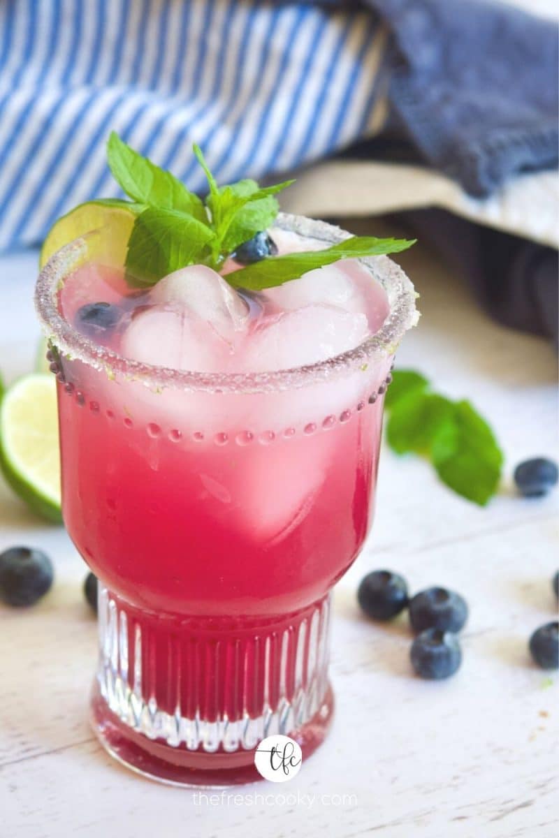 Close up image of a pretty decorative glass filled with blueberry limeade or lemonade, garnished with lime, mint and blueberries.