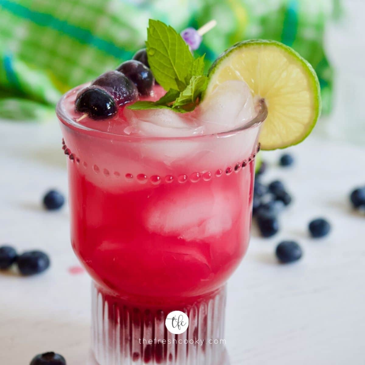 Image of a blueberry limeade drink in a pretty textured glass with a garnish of lime, mint and blueberries.