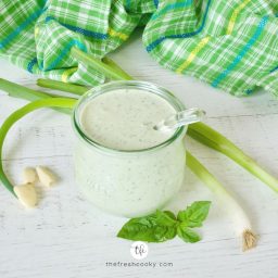 Square image of creamy buttermilk ranch dressing in a pretty glass jar with a glass ladle inside and fresh herbs and veggies laying around.