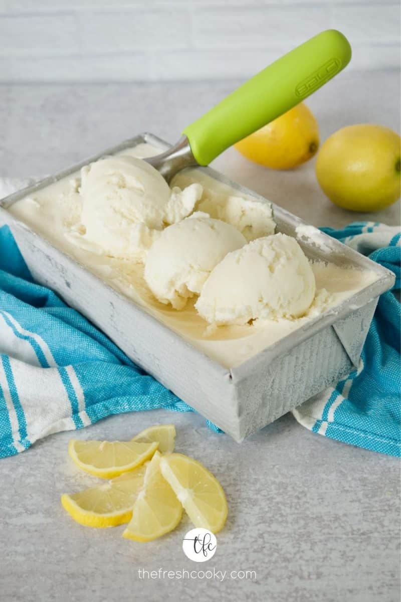 Pan filled with creamy lemon gelato ice cream with scoop and three scoops on top of the pan with lemons and blue tea towel behind.