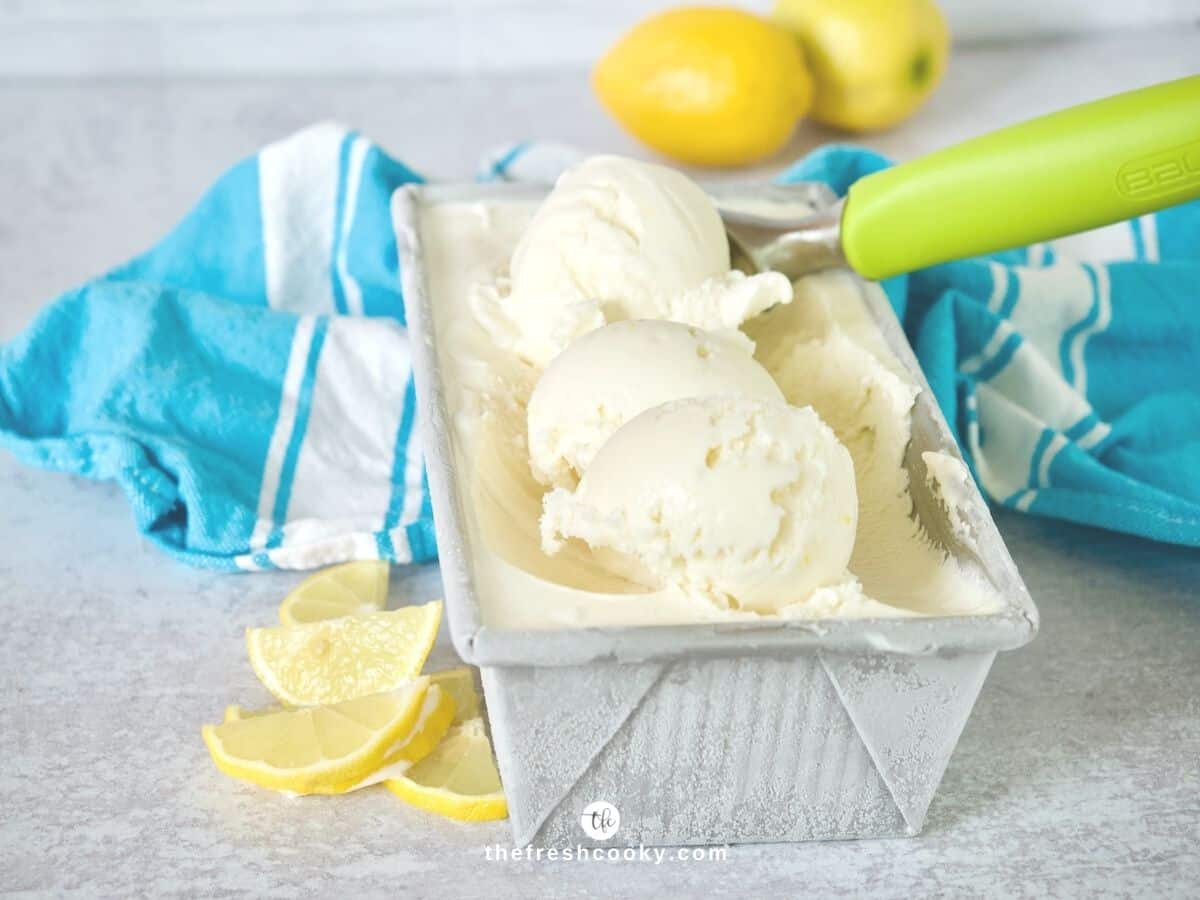 Creamy Lemon gelato ice cream in a loaf pan with an ice cream scoop and lemons behind.