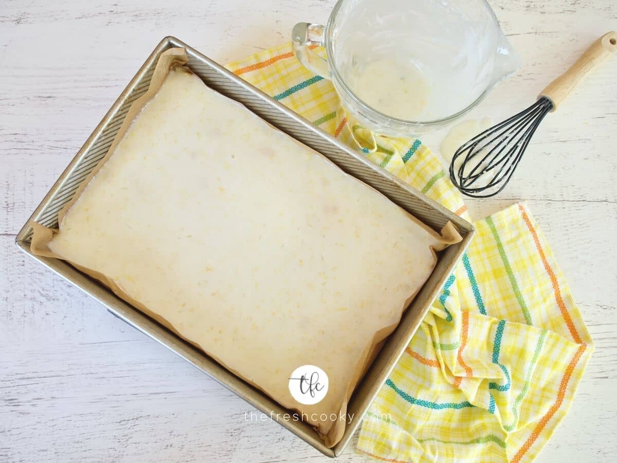 Top down image of glazed lemon bars in pan with whisk and bowl of glaze nearby.