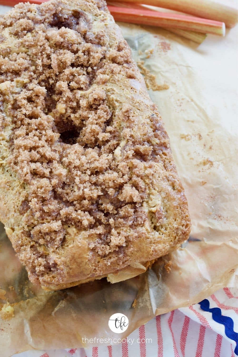 Loaf of Rhubarb bread with cinnamon crunch streusel topping sitting on parchment paper with rhubarb stalks behind.