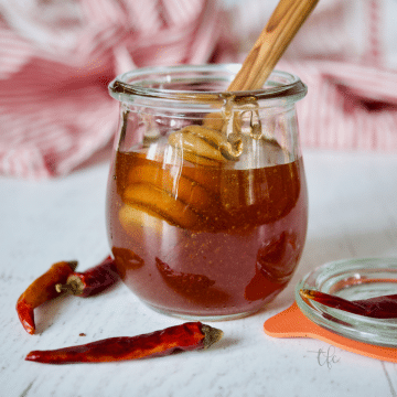 Square image of small jar of hot honey with chili peppers around and a honey server.