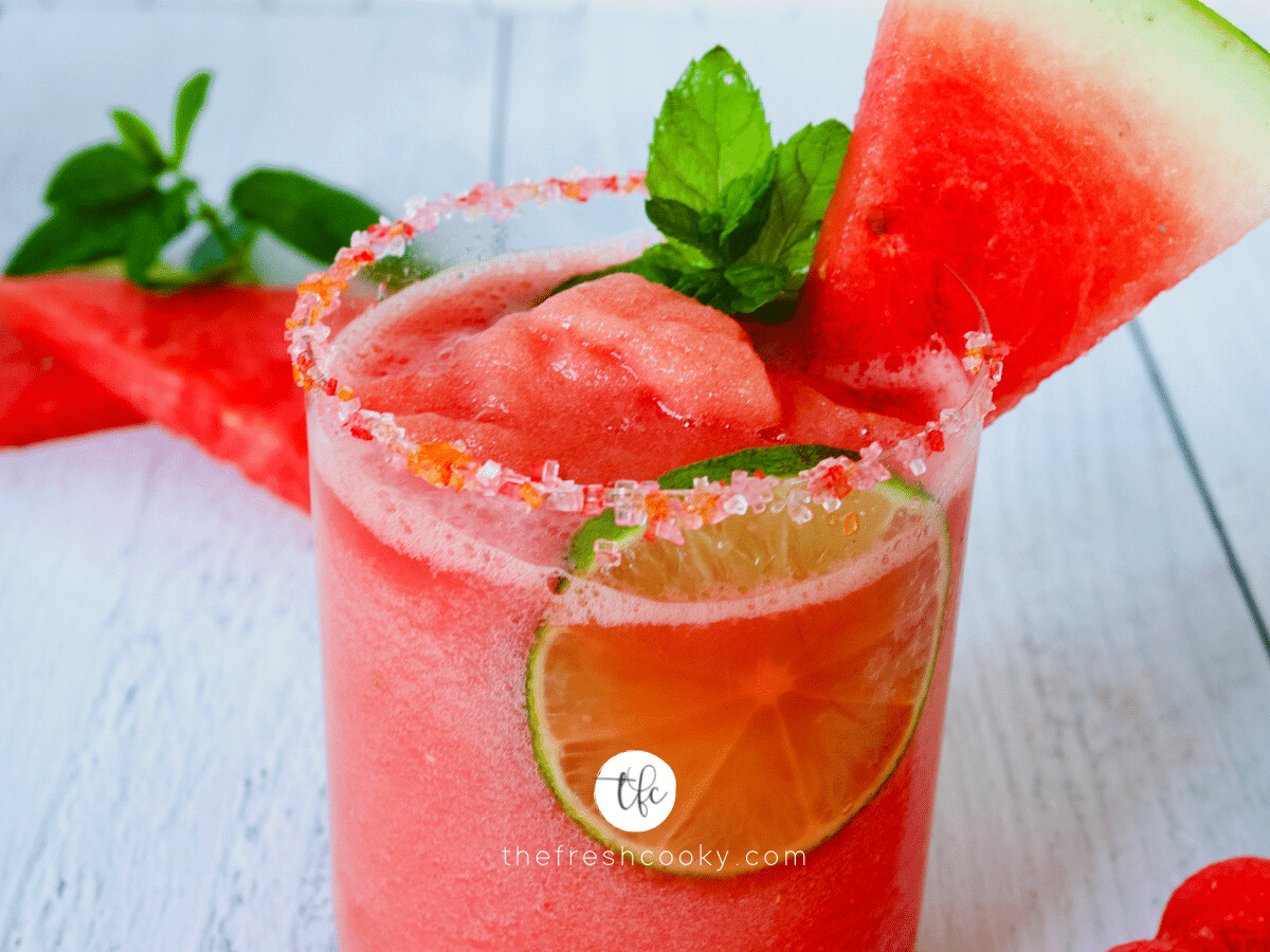 Brightly Colored Sugared Rim glass filled with bright red Watermelon Slush garnished with mint, lime and a wedge of watermelon.