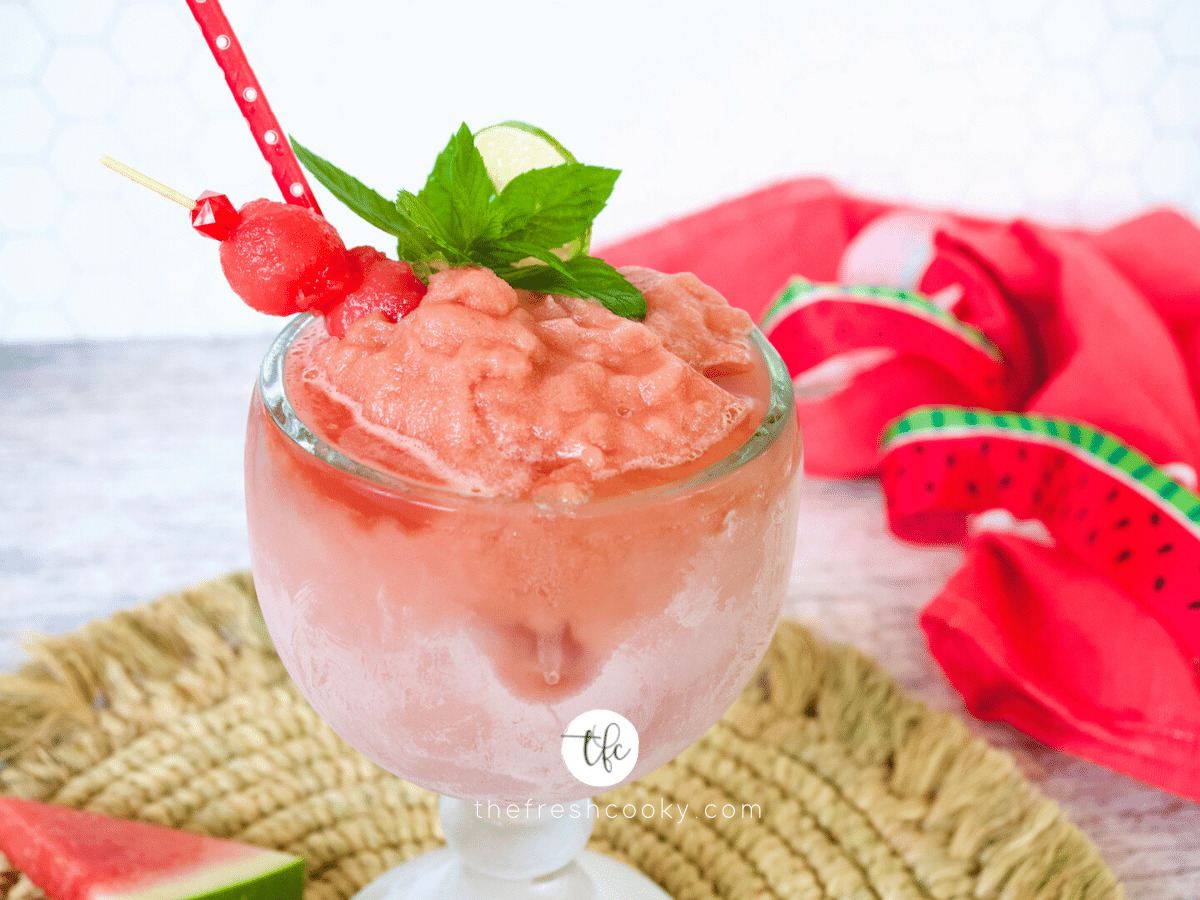 Schooner glass filled with watermelon slush garnished with melon balls, mint sprig and lime wedge.