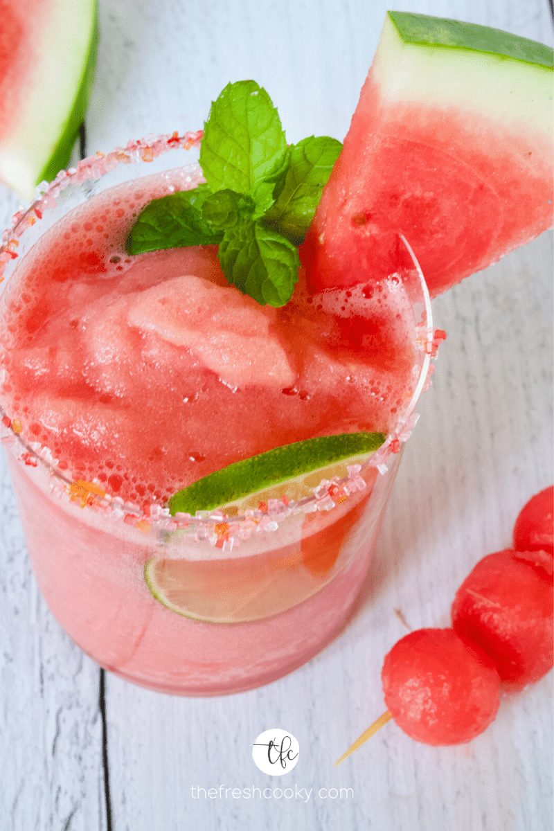 Glass of watermelon slush mocktail with lime wheel and fresh wedge of watermelon on a sugar rimmed glass.