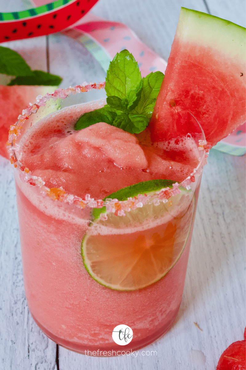 Sugar rimmed glass with watermelon slush inside, with lime wheel, mint sprig and wedge of watermelon.