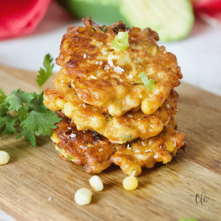 Zucchini Corn Fritters main image, three zucchini corn fritters stacked on one another with fresh veggies behind and cilantro beside.