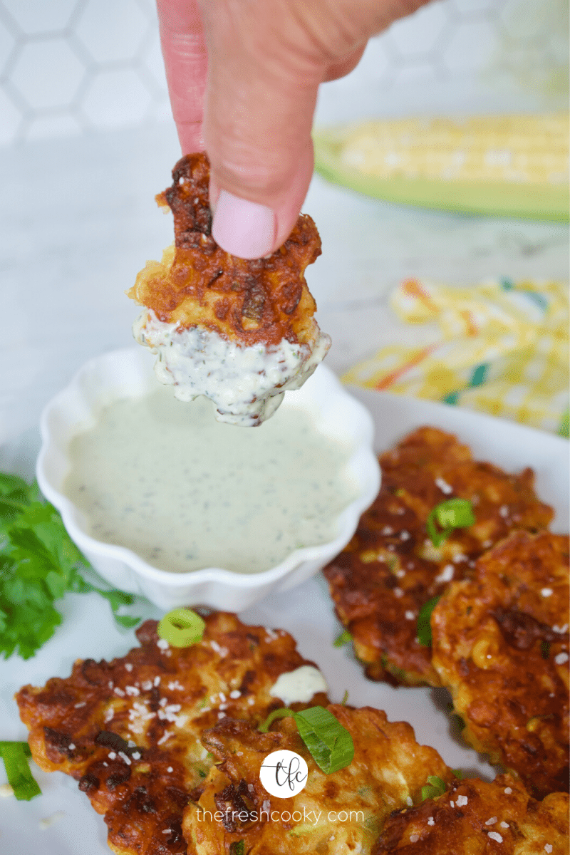 Hand dipping a zucchini corn fritter into some fresh ranch dressing with other fritters on the plate.