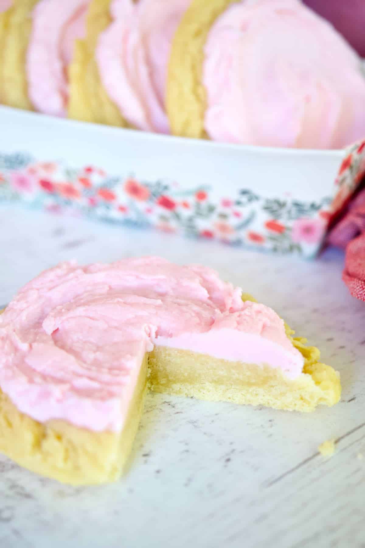 Crumbl Sugar Cookie on marble with pink frosting, with wedge removed revealing a soft, doughy, chewy center of the cookie.