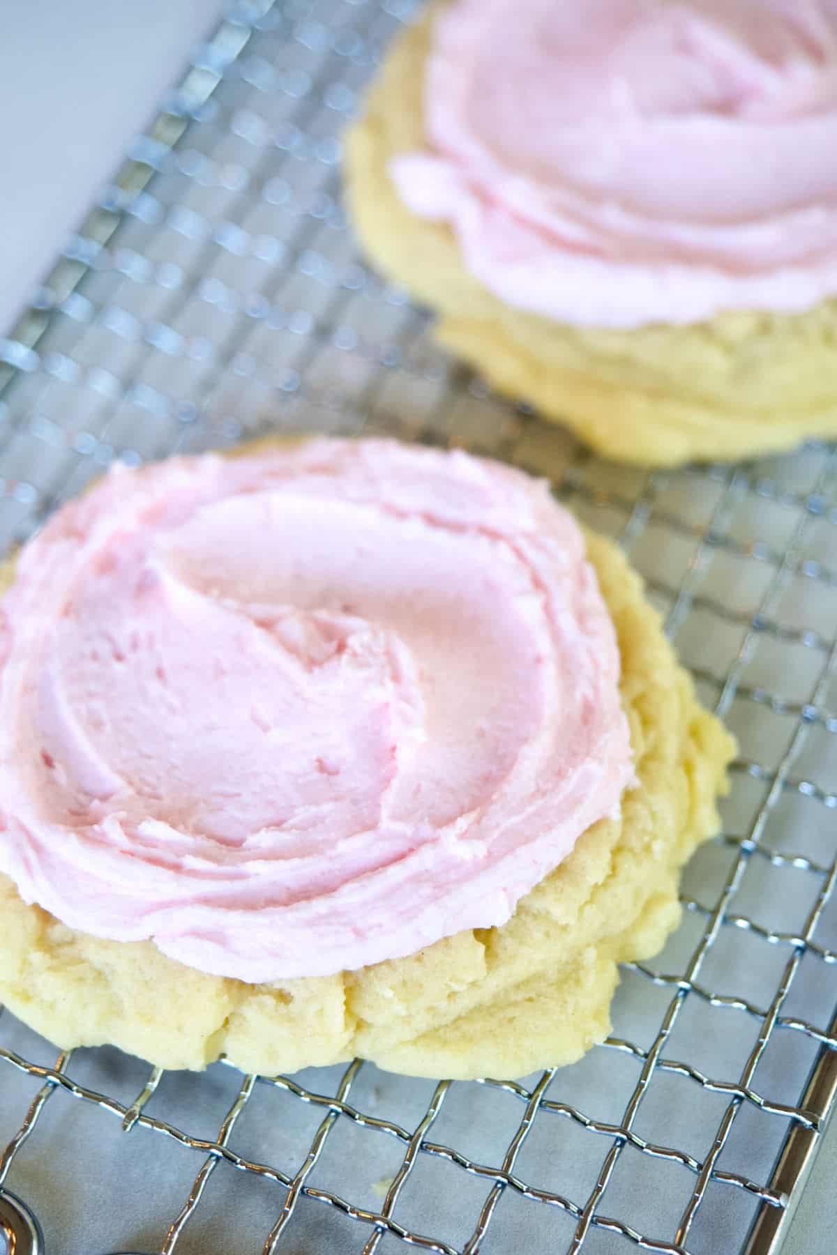 Easy Crumbl Sugar Cookies recipe with two frosted sugar cookies on a wire cooling rack.