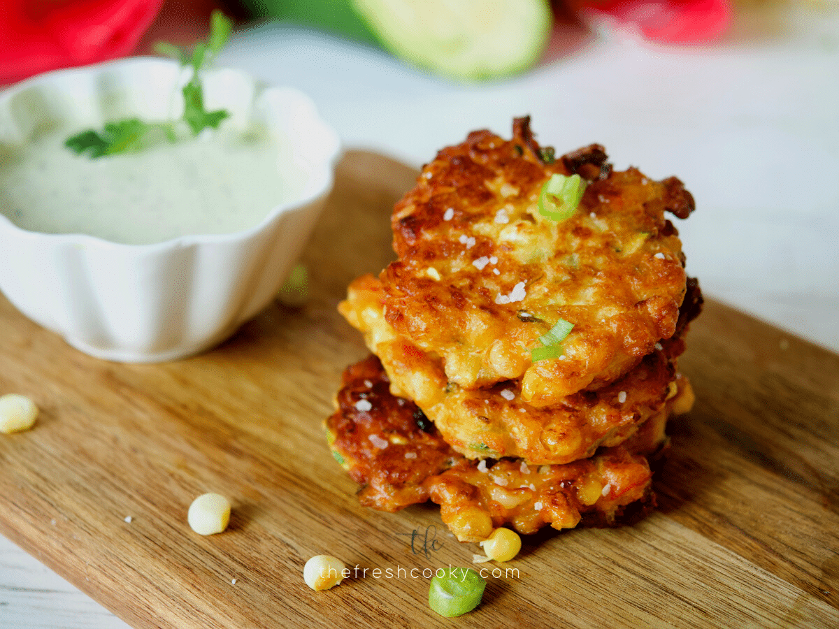 Delicious zucchini corn fritters stacked and sitting on a cutting board with some ranch dressing in a bowl behind.