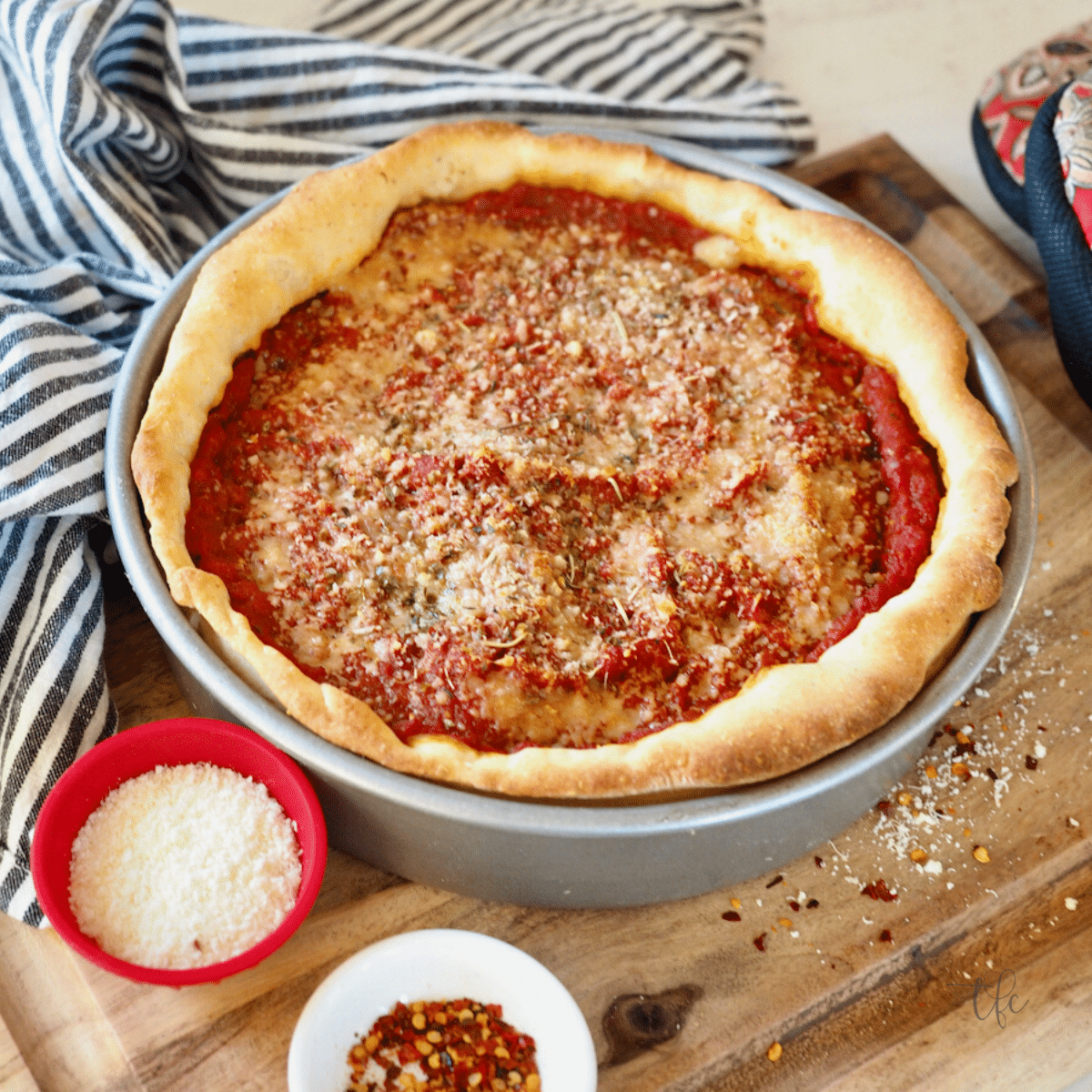 Top down shot of Chicago Style deep dish pizza in pan with parmesan cheese and red pepper flakes nearby.