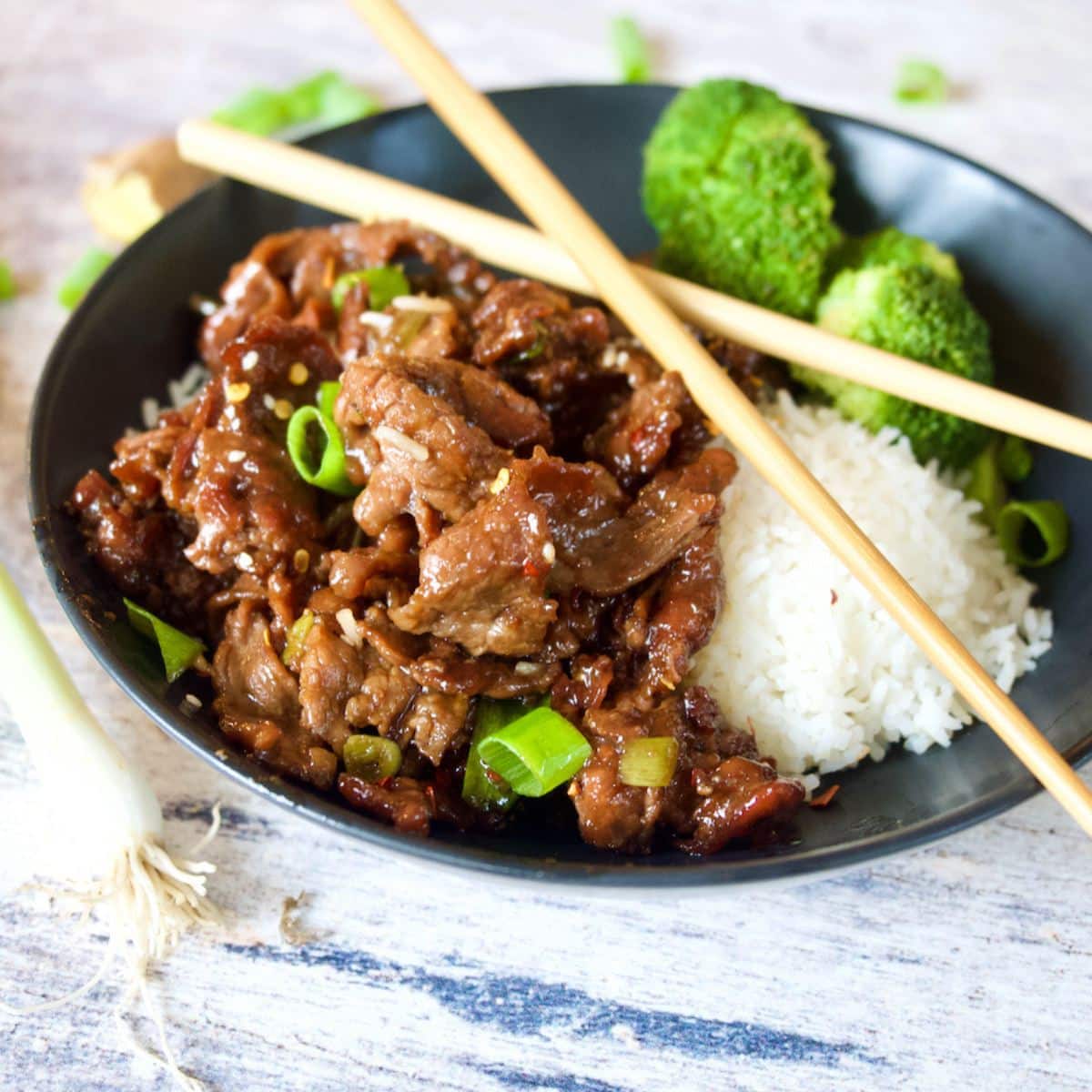 Mongolian Beef image of black bowl filled with Mongolian beef, rice and broccoli with chopsticks on top.
