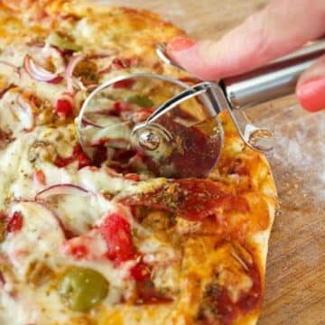 Cooked pizza on cutting board with hand using a pizza cutter to cut wedges of pizza.