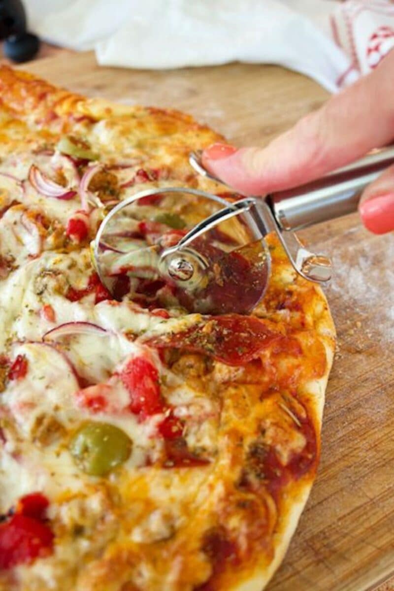 Cooked pizza on cutting board with hand using a pizza cutter to cut wedges of pizza.
