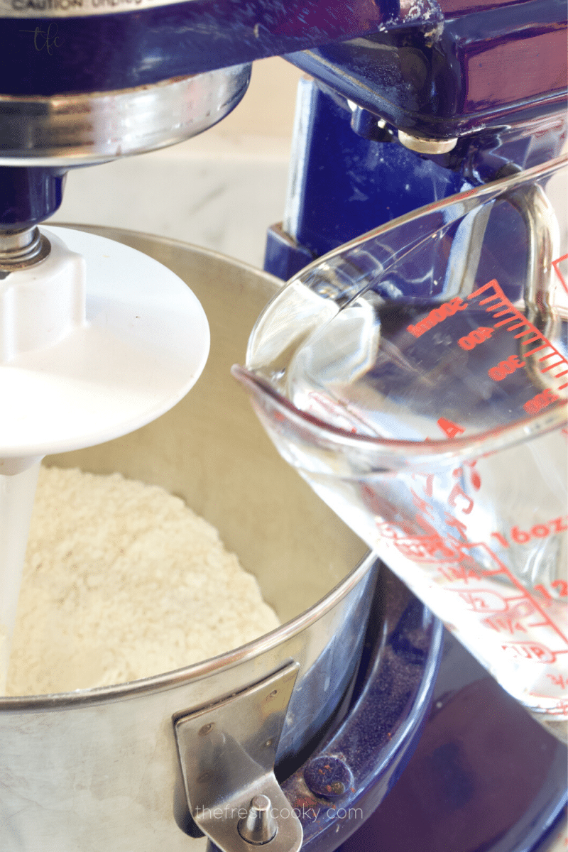 Pouring water into stand mixer bowl filled with yeast, cornmeal, sugar and flour to make deep dish butter crust pizza dough. 