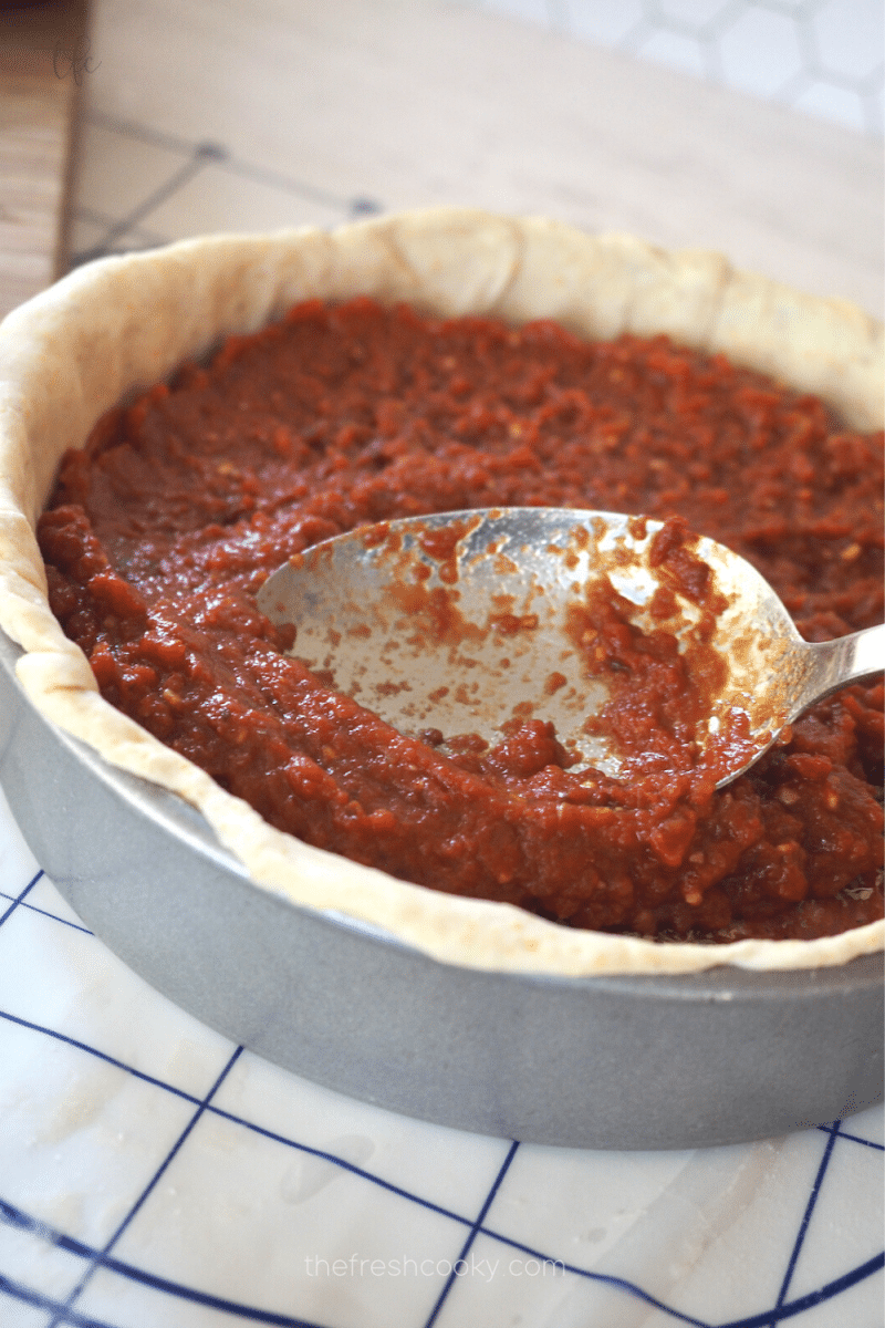 Spreading chunk tomato sauce on top of deep dish pizza toppings. 