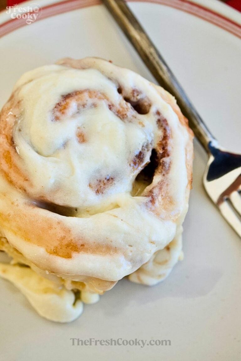 A close-up shot of a soft, gooey cinnamon roll smothered in cream cheese frosting, on a white plate with a rustic fork. To pin.