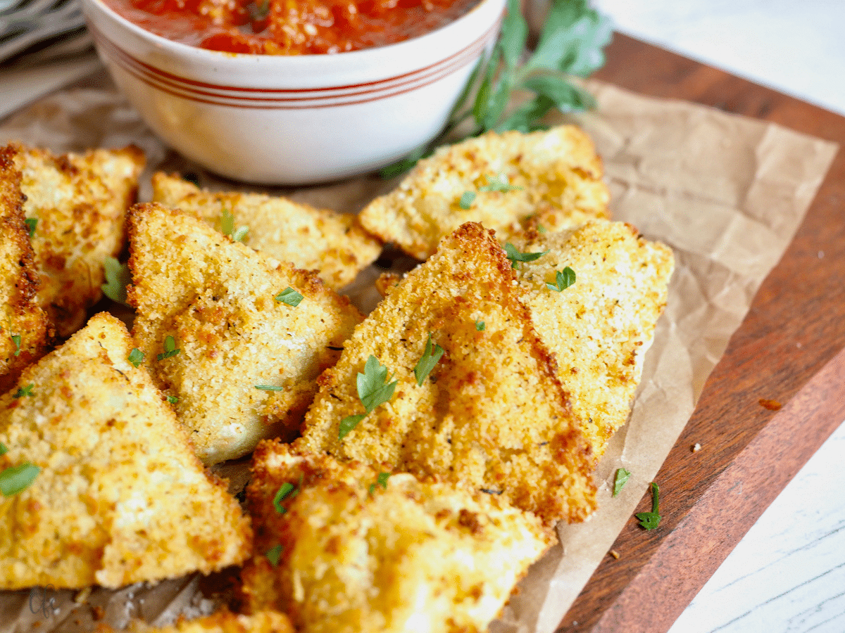 Air fryer ravioli on board with marinara sauce behind.