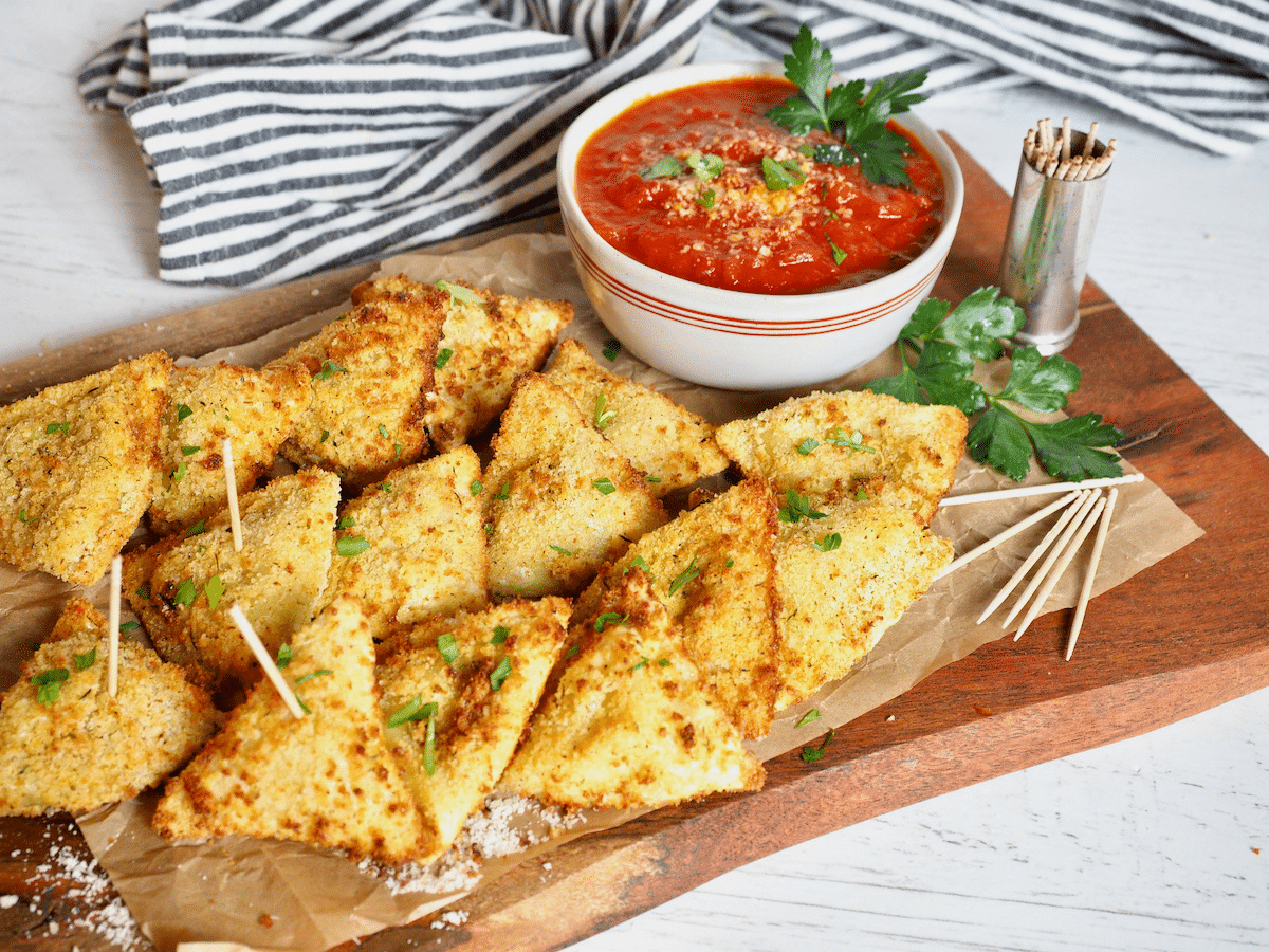 Air fryer ravioli top down shot with raviolis on a cutting board with toothpicks and a bowl of marinara sauce.