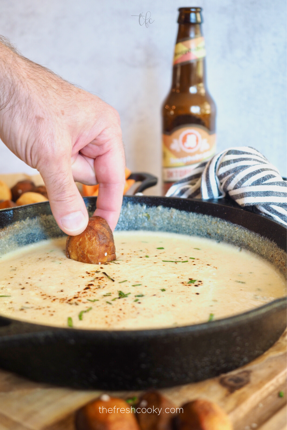 Easy Beer Cheese Dip with pretzel dip in cast iron skillet and mans hand dipping a pretzel bite into dip with a bottle of beer behind.