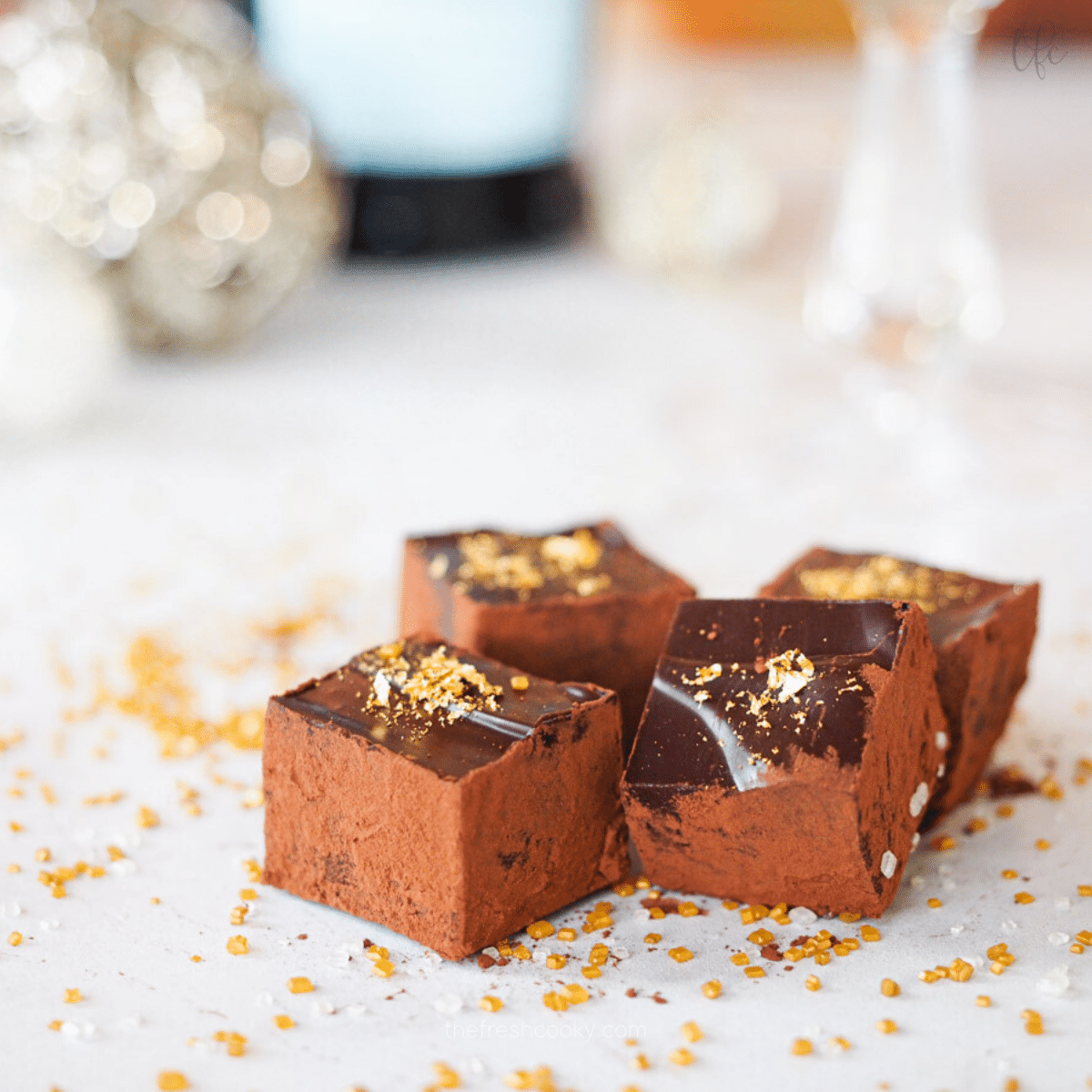 Square image of 4 Champagne Truffles dusted in cocoa powder and decorated with gold leaf sitting on marble with bottle of champagne behind and a stemmed glass.
