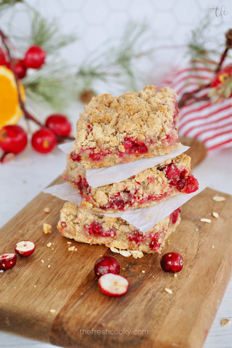 Easy Cranberry Crumble Crunch Bars with Oatmeal and orange stacked three on top of the other, on a pretty wooden cutting board with orange and greens in background.