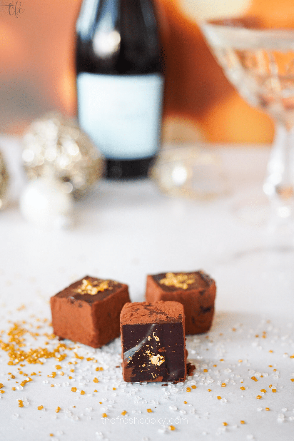 Champagne Truffles three of them with gold leaf, on marble with sprinkles around and a bottle and glass of champagne in background.