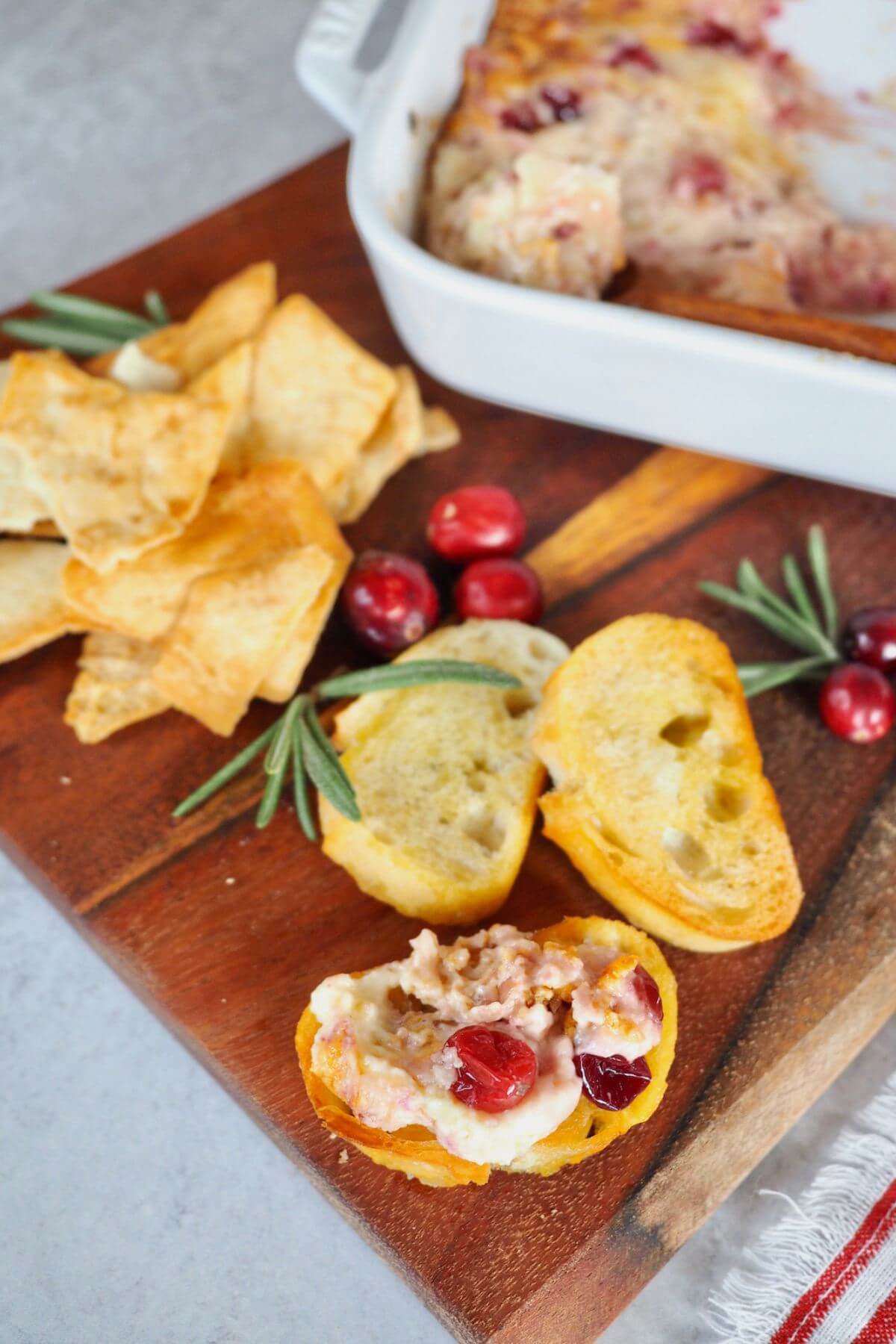 Cranberry cream cheese dip smeared on crostini next to fresh herbs and cranberries.